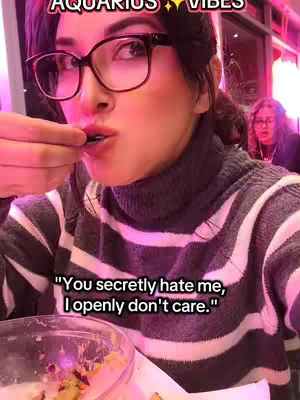 A woman with dark hair and glasses is eating from a bowl. She is wearing a striped gray and white sweater. In the background, another woman is visible.