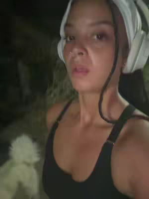 A selfie-style shot featuring a woman in a dark-colored tank top. She is wearing a white head covering and large white headphones. The background is blurry and dark.