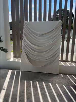 An outdoor shot featuring a large, off-white plaster artwork. The artwork is draped with fabric, creating soft, curved lines, suggestive of drapery. It's positioned on a concrete surface with sunlight casting long, parallel shadows from a fence, creating a contrast between light and shadow.
