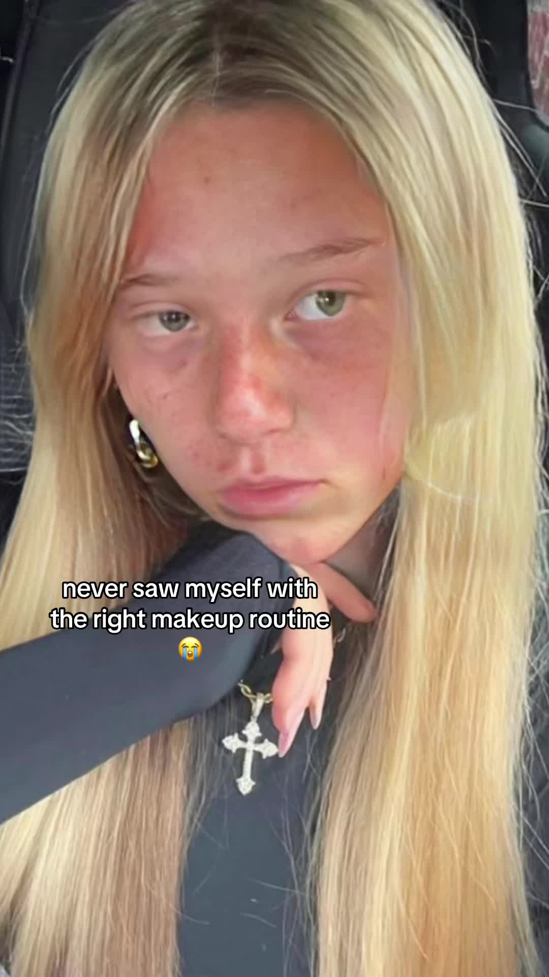 The image is a selfie-style shot featuring a person with long blonde hair, seen in a close-up view. The person is positioned slightly off-center, with their face angled towards the camera. The background is blurred, suggesting an indoor setting, possibly a car. Overlayed text at the bottom reads 'never saw myself with the right makeup routine' accompanied by a crying emoji.