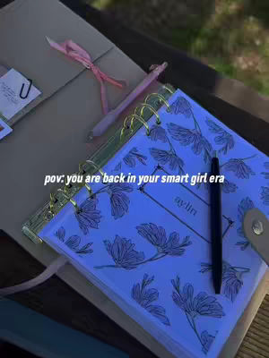 The cover image shows a notebook with a floral design resting on a surface, likely a wooden bench. The notebook is open to display a blank page with the name "aylin" written on it. A pen is placed near the notebook. In the background, a pen, pink ribbons, and other objects can be seen. The image is lit with a cool, blueish tint.