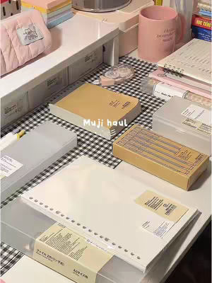 The image showcases a well-lit, aesthetically arranged collection of Muji stationery items on a white surface. Various Muji notebooks, clear boxes, and other stationary accessories are neatly organized. The background features a black and white checkered pattern, and a pink mug is partially visible, adding to the scene's appeal.