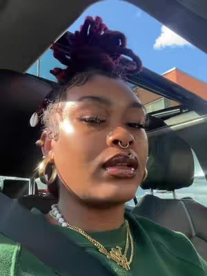 A selfie-style video shows a woman inside a car, likely a sunny day. Her face is prominent, with a nose ring, full lips, and styled hair. She wears a green top and gold jewelry, including hoop earrings and a necklace. She is looking towards the camera. The car interior and sky through the sunroof are visible in the background.