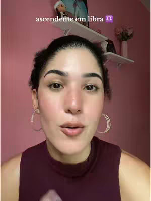 A woman with dark hair and hoop earrings looks directly at the camera. Above her, a text overlay reads 'ascendente em libra,' with the Libra symbol next to it. The background is a pink wall, and a shelf behind the woman holds decorative items.