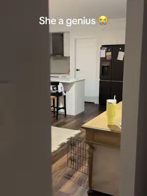 View from a doorway into a kitchen. A yellow cup with a straw sits on a table. The kitchen has white cabinets and a black refrigerator. Text 'She a genius' is added at the top of the frame.