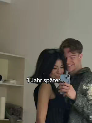 A selfie-style shot of a smiling couple. The woman, with dark hair, is holding a phone, partially obscuring her face. The man is smiling broadly, with his arm around her. The background is a plain, neutral-colored wall.