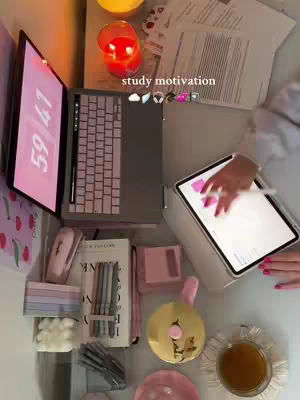 A top-down shot of a study setup. A laptop screen shows a pink digital clock, with a keyboard set in front of it, a tablet with stylus, alongside stationary, candle, and a cup of tea on a desk. A hand touches the tablet in the foreground.