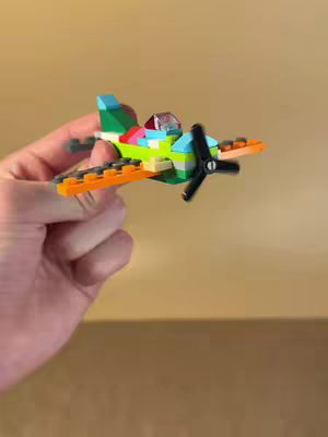 A hand holding a small, multicolor toy airplane made of interlocking plastic bricks. The plane has a green fuselage, blue and red accents, orange wings, and a black propeller.