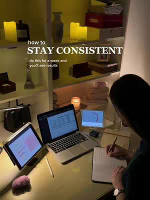 A softly lit workspace features a woman working on a laptop and a tablet. The scene includes shelves with decor and warm lighting. The text overlay reads, 'How to STAY CONSISTENT,' with an additional line, 'do this for a week and you'll see results.'