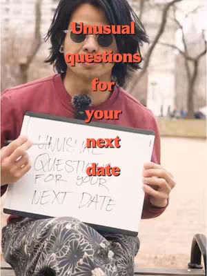 A person with sunglasses holds up a white board with text. The background is an outdoor public space.