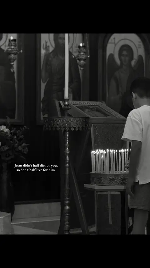 A black and white photo shows the interior of a church. A young man stands in front of a display of lit candles, with his back to the camera.  There are religious icons on the wall and lit candles.  The text overlay states: "Jesus didn't half die for you, so don't half live for him."