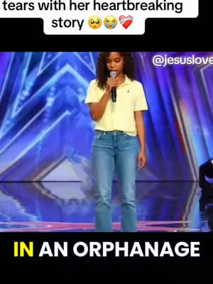 The image shows a young woman standing on a stage holding a microphone, bathed in the glow of stage lights. She's wearing a yellow polo shirt and blue jeans. The background is a blurred display of blue and purple lights. Text overlays include emotional emojis and the phrase 'IN AN ORPHANAGE.'