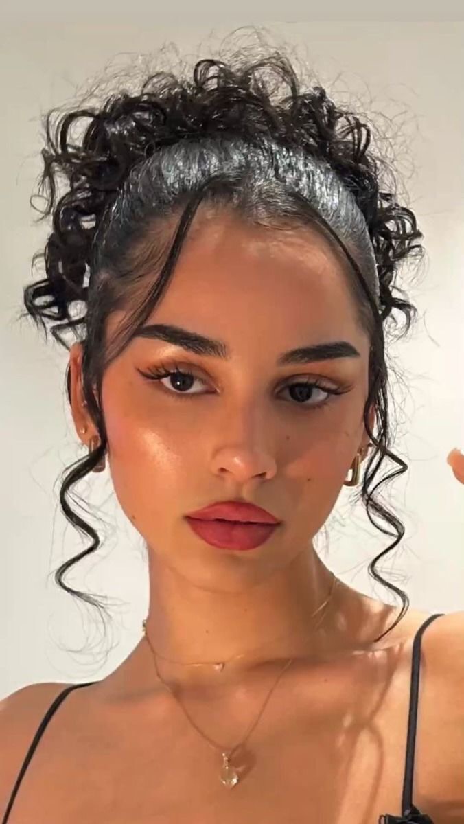 A close-up shot of a woman with dark curly hair pulled back in a high bun and delicate face. She wears gold hoop earrings and a minimal black top, and shows a slight expression with red lipstick, perfect eyebrows, and long eyelashes add to her glamorous look. The focus is on her facial features, with light reflecting off her skin creating a soft, glowing effect.