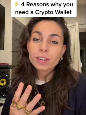 A woman is centered in the frame, looking directly at the camera with a slight smile and open mouth, suggesting she is speaking. She has long, dark brown hair. Her left hand is up, displaying her fingers, with several gold rings. A text overlay at the top reads, "4 Reasons why you need a Crypto Wallet". The background includes elements of a room interior, such as curtains and a dark object resembling equipment.