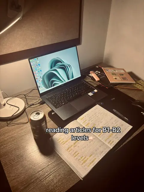 A dimly lit desk contains a laptop displaying a teal abstract design on the screen, an open notebook with handwritten notes, a can of dark soda, and other study materials. The scene suggests a study session or work environment.