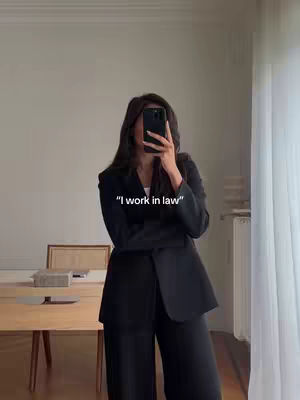 A woman in a black suit takes a mirror selfie, obscuring her face with her phone. She stands in a well-lit room; in the background are a table, chair, and curtains.