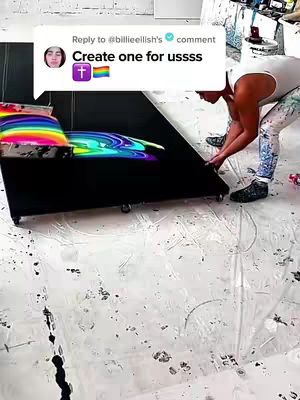 A person holding a canvas where wet paint in bright colors of the rainbow is spread on a black surface, on smooth white floor, with a speech bubble from billieeilish containing text and a rainbow flag icon.