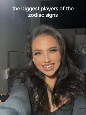 A selfie-style shot featuring a woman with dark hair and makeup, smiling. Text overlay at the top reads 'the biggest players of the zodiac signs'. The background includes a glimpse of a room.