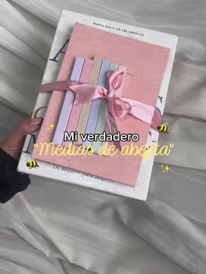 The image features a book with a pink cover, likely a journal or a Bible, adorned with a pink ribbon and several colorful bookmarks tied to it. The book rests on another book, possibly a larger Bible. The composition showcases a soft color palette dominated by pinks and pastels, with added decorative elements such as cute bee stickers and text overlays.