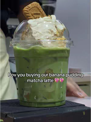 A close-up shot of a matcha latte in a clear plastic cup with a dome lid. The drink has a vibrant green hue, and is topped with whipped cream and a cookie. The cup sits on a dark surface, with a blurred background.