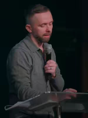 A man with short, light-colored hair and a trimmed beard speaks into a microphone behind a podium. He's wearing a button-down shirt with a collar. The background is dark and out of focus.