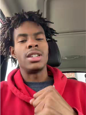 A person inside a vehicle looks directly at the camera, wearing a red hoodie. The background includes portions of the car's interior and the view outside the window.