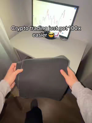The cover image shows the first-person perspective of a person holding a black pad-like object against their chest. The background includes a computer monitor displaying a stock chart and a white desk. The person's hands and arms are visible as they hold the object, which partially obscures their body and the lower portion of the screen. The text overlay reads 'Crypto trading just got 100x easier' with an emoji depicting sunglasses and an upward trending graph.