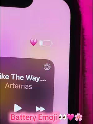 The image shows a close-up of an iPhone screen, displaying the battery status with added emoji customizations. In the top portion of the screen, a pink heart emoji is positioned next to the battery icon. The lower area showcases an Apple Music interface with the song "Like The Way..." by Artemas playing, and the text "Battery Emoji" with various emojis is added at the bottom.