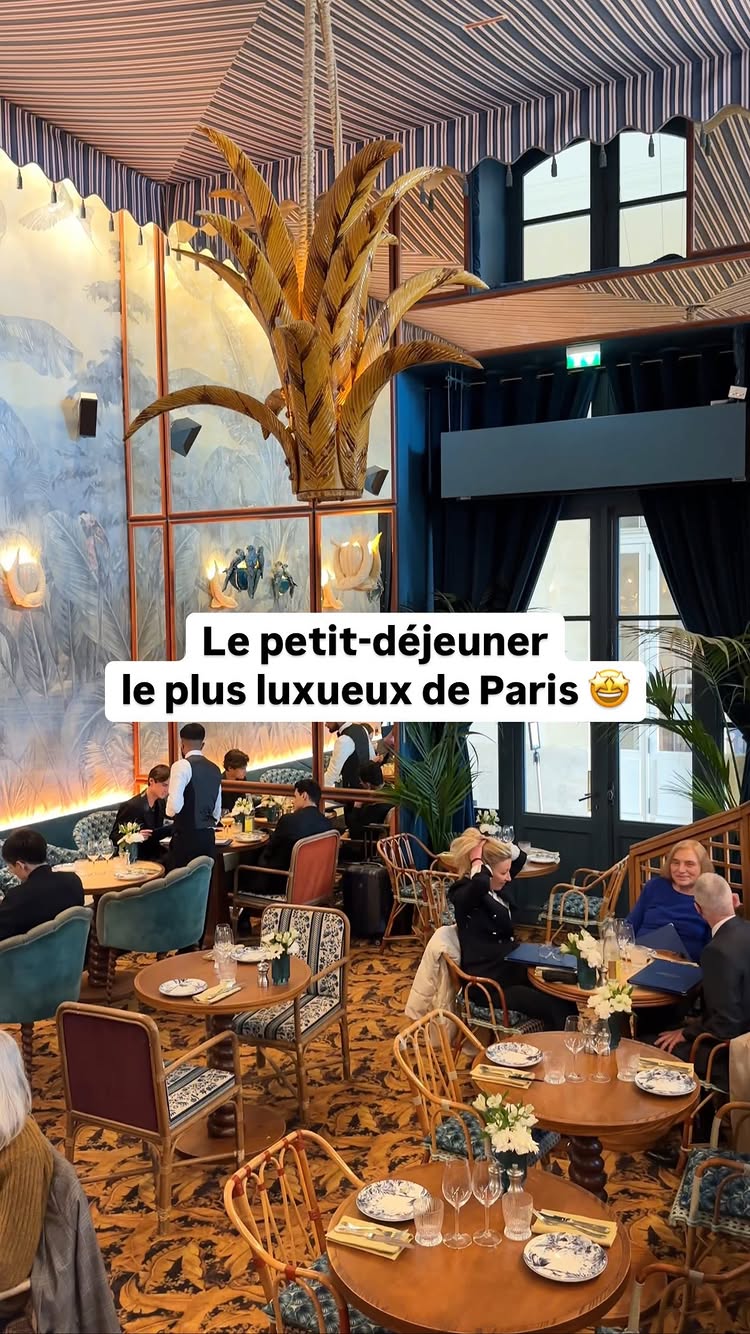 The image showcases a luxurious restaurant interior. A gold-colored palm tree-shaped chandelier hangs from a striped blue and red awning ceiling. Tables with food and people are visible, including a waiter. The setting is opulent, displaying a mix of modern and classic decor.
