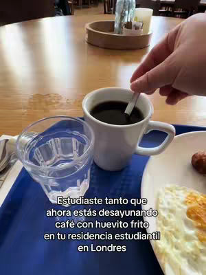 A photo of a breakfast spread on a blue tray in a bright, neutral-lit setting. A white mug of dark coffee sits beside a clear glass partially filled with water. A plate on the right holds a fried egg and a sausage. The background includes a wooden table and a few other items slightly out of focus.