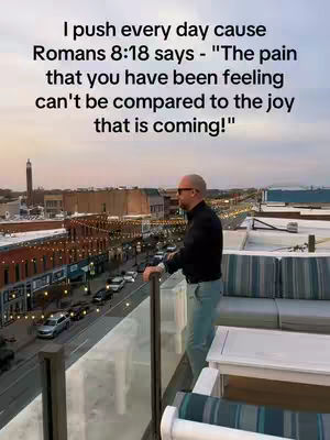 A man stands on a rooftop overlooking a city at dusk. Text overlays the image with a quote from Romans 8:18 about enduring pain for future joy.