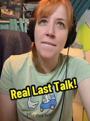 A woman with red hair and a green shirt is looking upwards with a slight bite on her lower lip, headphones around her neck. The image is a selfie.