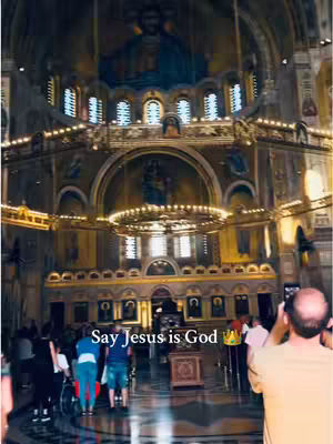 Interior shot of a large church with people present. The ceiling is high with decorative elements including paintings and ornate lighting. A person is taking a photo, included as a main figure in the frame.