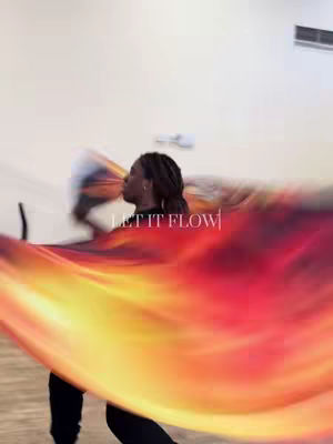 A woman is dancing with a large, colorful flag that has an orange and red flame-like design on it. The background has a light wall.