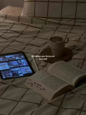 A cozy, dimly lit scene. A bed with a patterned comforter and pillows dominates the frame. On the bed, there's an open book, a tablet displaying an interface, and a cup of coffee on a wooden coaster. The overall tone is warm and inviting.