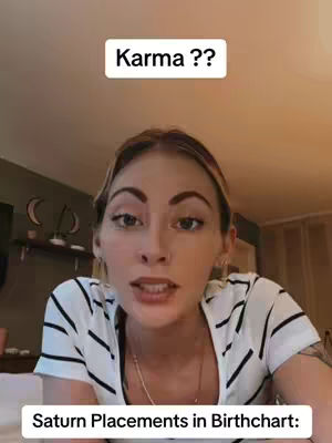 A woman with blonde hair and a white and black striped shirt is in front of a camera. The video has two text overlay boxes with question and answer prompts about karma and Saturn placements in a birth chart. The background includes a wall, some decor, and lighting fixtures.