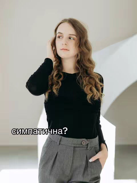 A young woman with long, wavy brown hair is the focus of the image. She's wearing a black long-sleeved shirt and gray pleated trousers. Her left hand is in her pocket and her right hand is touching her hair. The background is a soft, blurred gradient of light colors