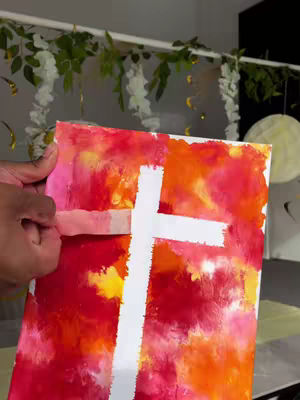 A hand holds a painted canvas with a white cross on a multicolor background, and is peeling off some tape.