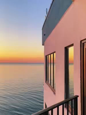 The image shows a serene sunset over the ocean. A building with a white exterior and teal roof is on the right. The sky transitions from blue to a warm gradient of oranges and yellows, reflecting on the water's surface.
