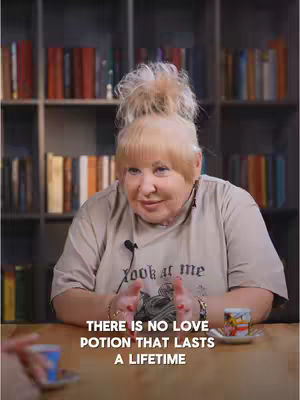 A woman with blonde hair in a bun sits at a wooden table with two small cups and saucers, gesturing with both hands. She wears a light-colored t-shirt with 'look at me' printed on it. A bookshelf filled with books is in the background. Text at the bottom reads, 'THERE IS NO LOVE POTION THAT LASTS A LIFETIME'.