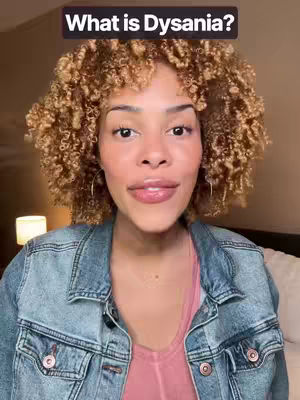 A medium shot of a woman with curly brown hair and a denim jacket, centered on her face. Text overlay at the top reads "What is Dysania?" The woman has fair skin, a pink top underneath, and is looking directly at the camera with a slight smile. The background is blurred and warm-toned.