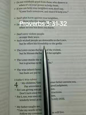 A close-up image shows an open book on a textured surface. In the center, a silver pen points downward, aligned with a specific line of text. A text overlay reads "Proverbs 3:31-32" in a clear, sans-serif font, positioned above the relevant text passage. The background shows the printed pages of the book, with clear, readable text.