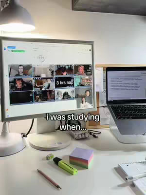 A desk setup with a computer monitor displaying several video call windows and a laptop in the background. There are also office supplies on the desk such as a highlighter, sticky notes, and a pencil under a lamp.