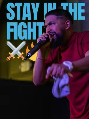 A man with a beard and a red shirt is holding a microphone and pointing with his finger, with text overlayed that says, "Stay in the fight". There are two swords and an image of flames in the background. The background in general is dark, making the person in the image more prominent.