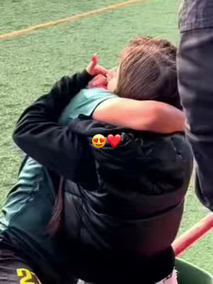 Two individuals are captured in a tight embrace, their faces close. The background is a green field with sidelines visible. The image features two emoji stickers.