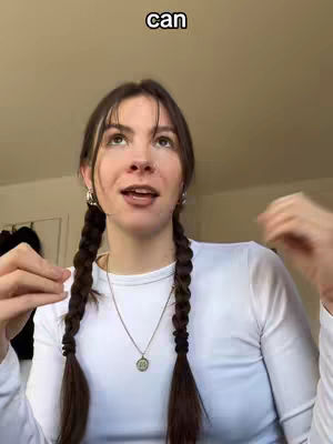 A young woman with long brown hair styled in two braids is looking upwards with an expression of curiosity, her hands gesturing around her head. The background is a plain, off-white wall, possibly indoors. She wears a white long-sleeved shirt and a pendant necklace. The word 'can' is overlaid in the image near the top.