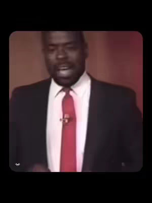 A man in a suit and tie is centered in the frame, speaking. The background is a blurred maroon color. The man is black and wearing a black suit with a white shirt and a red tie. A microphone is clipped near the tie. The image is a rounded rectangle with a black background.