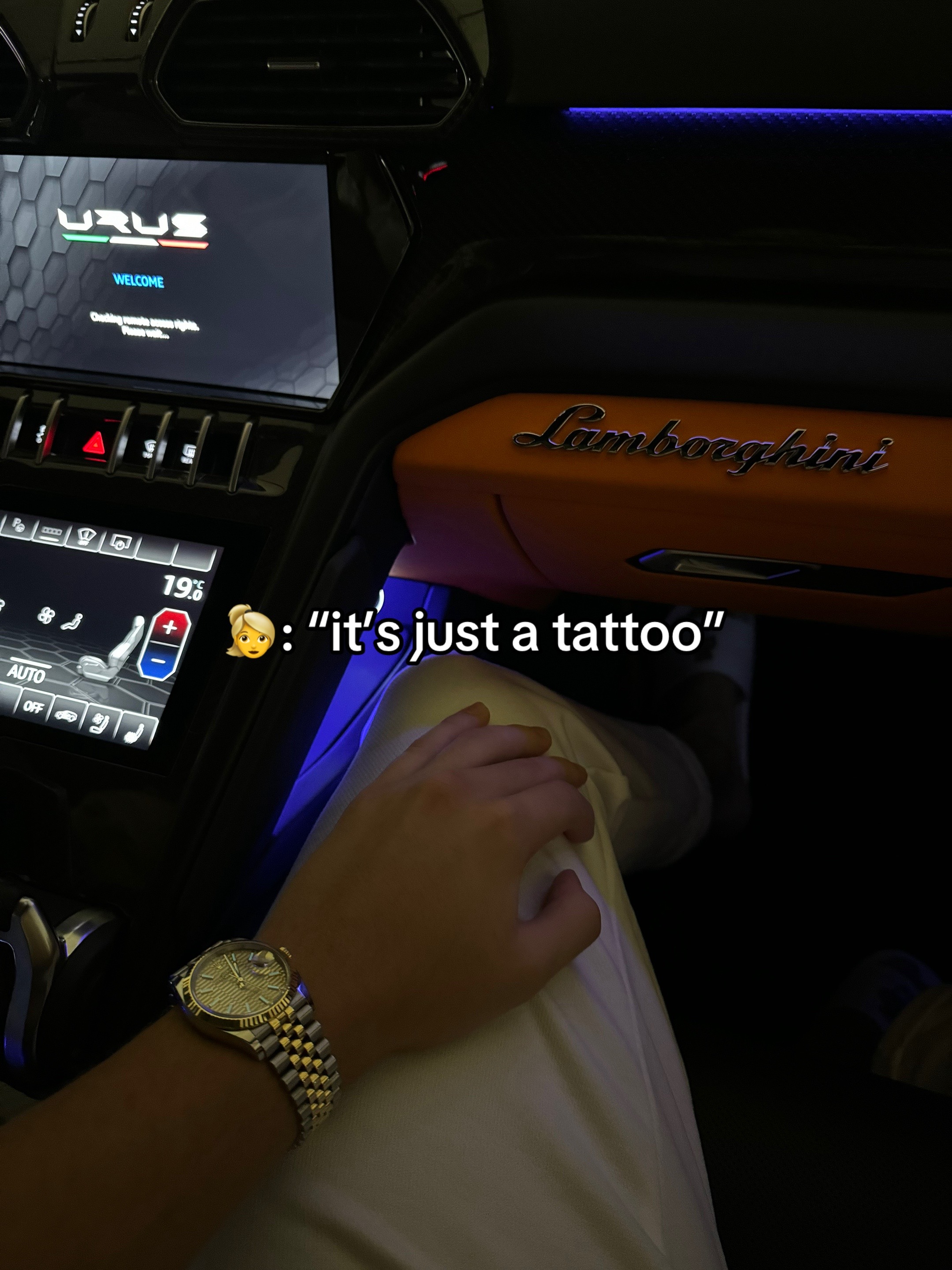 The image shows the interior of a Lamborghini, with the dashboard visible and a person's arm and wrist in the foreground. A hand with a watch is resting in the person's lap. Text overlay includes an emoji of a woman and the phrase "it's just a tattoo."
