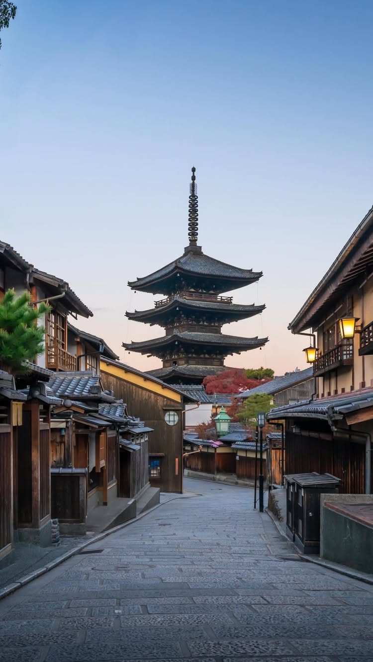 A photograph of a stone street in a Japanese neighborhood, with dark wooden buildings on either side and a tall pagoda in the background. The sky is a gradient of blue and pink.