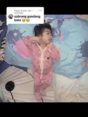 The image shows a young child lying down in a bed, wearing a pink onesie. The child is resting on a blue and white patterned pillow. The background includes a part of a bed sheet and a comment bubble overlaying the image.